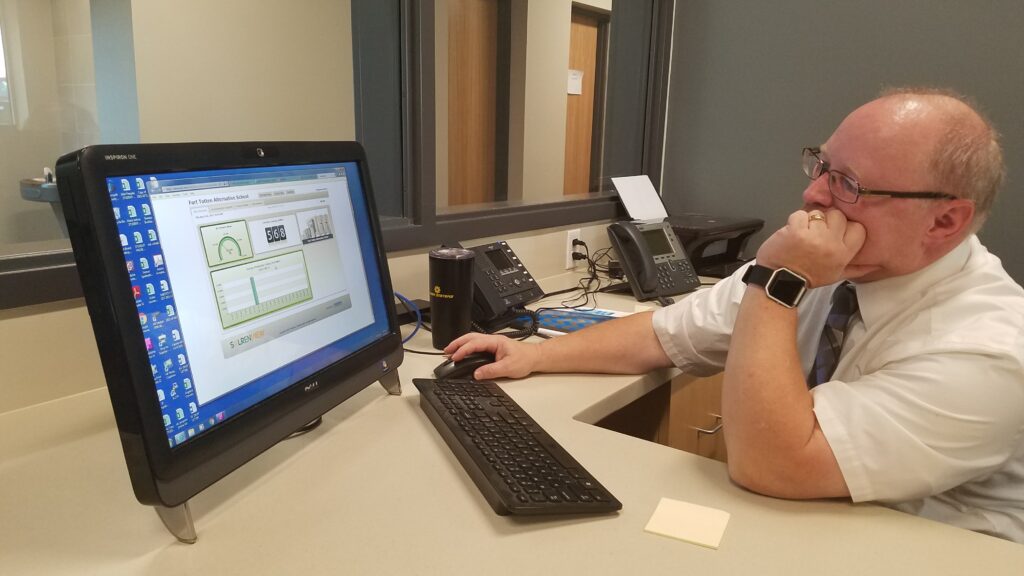 Man in white shirt and glasses sits at a desk, looking at a monitor displaying a dashboard with charts and graphs in an office setting.