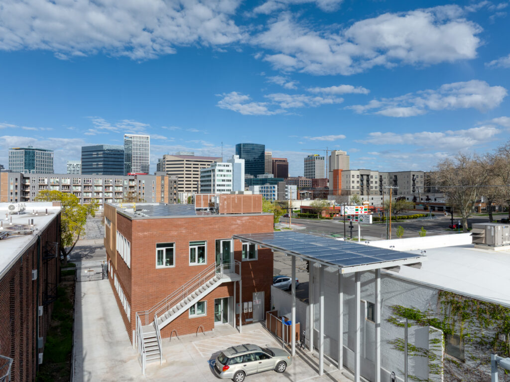 Rooftop view of brick buildings and a parking area with the city skyline in the distance under a blue sky with clouds: urban, daytime scene.