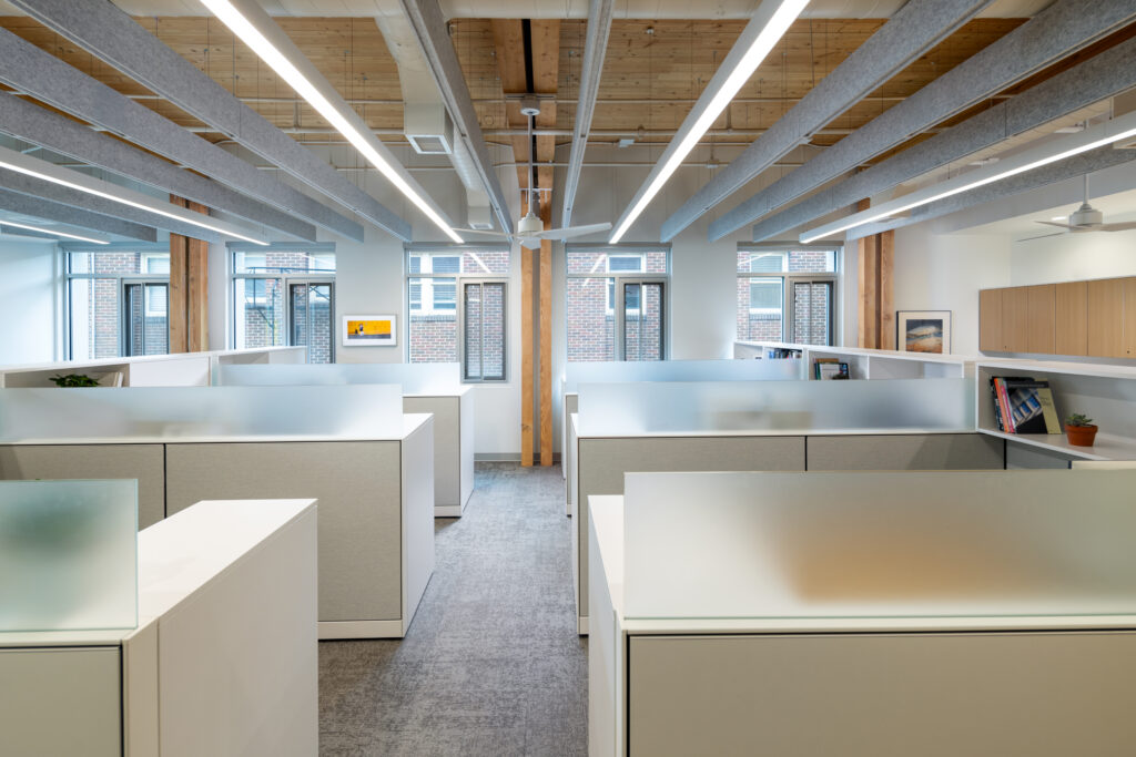 Open-plan office with white cubicles, frosted glass dividers, and exposed wood ceiling beams.
