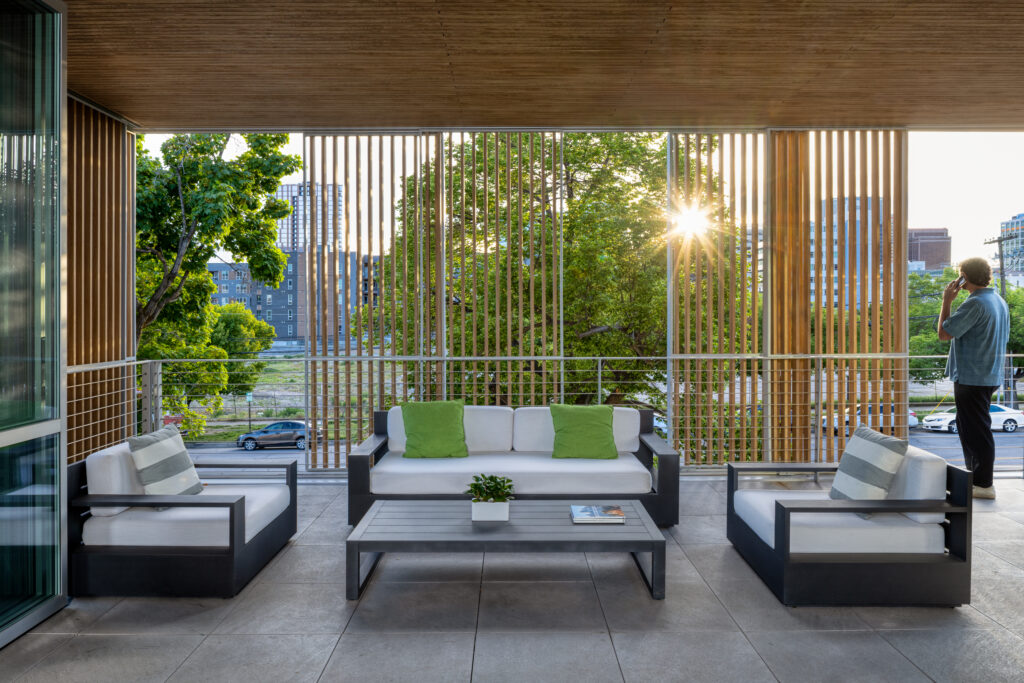Outdoor balcony lounge with white-cushioned sectional sofas and green pillows, wooden slat screens, and a sunset behind trees in the cityscape.