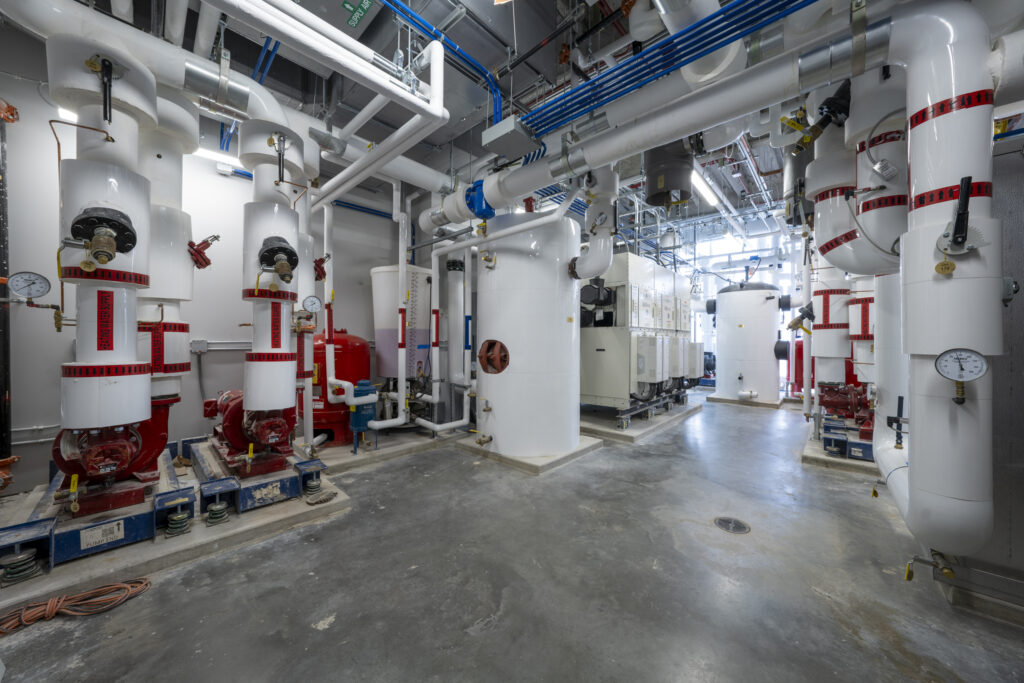 Industrial mechanical room with large white insulated tanks, red-striped valves, gauges, and a maze of white and blue pipes overhead.
