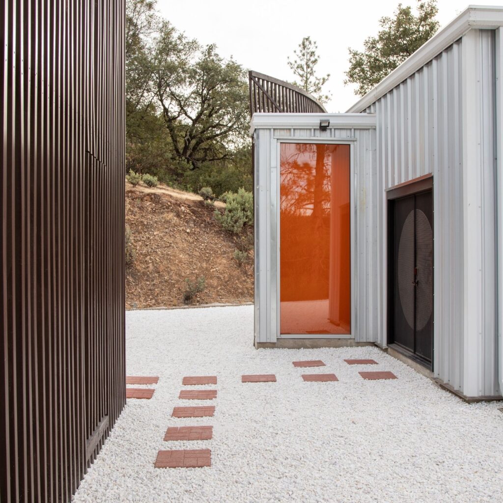Narrow courtyard between dark brown slatted wall and metal-clad building, with a stepping-stone path on white gravel.