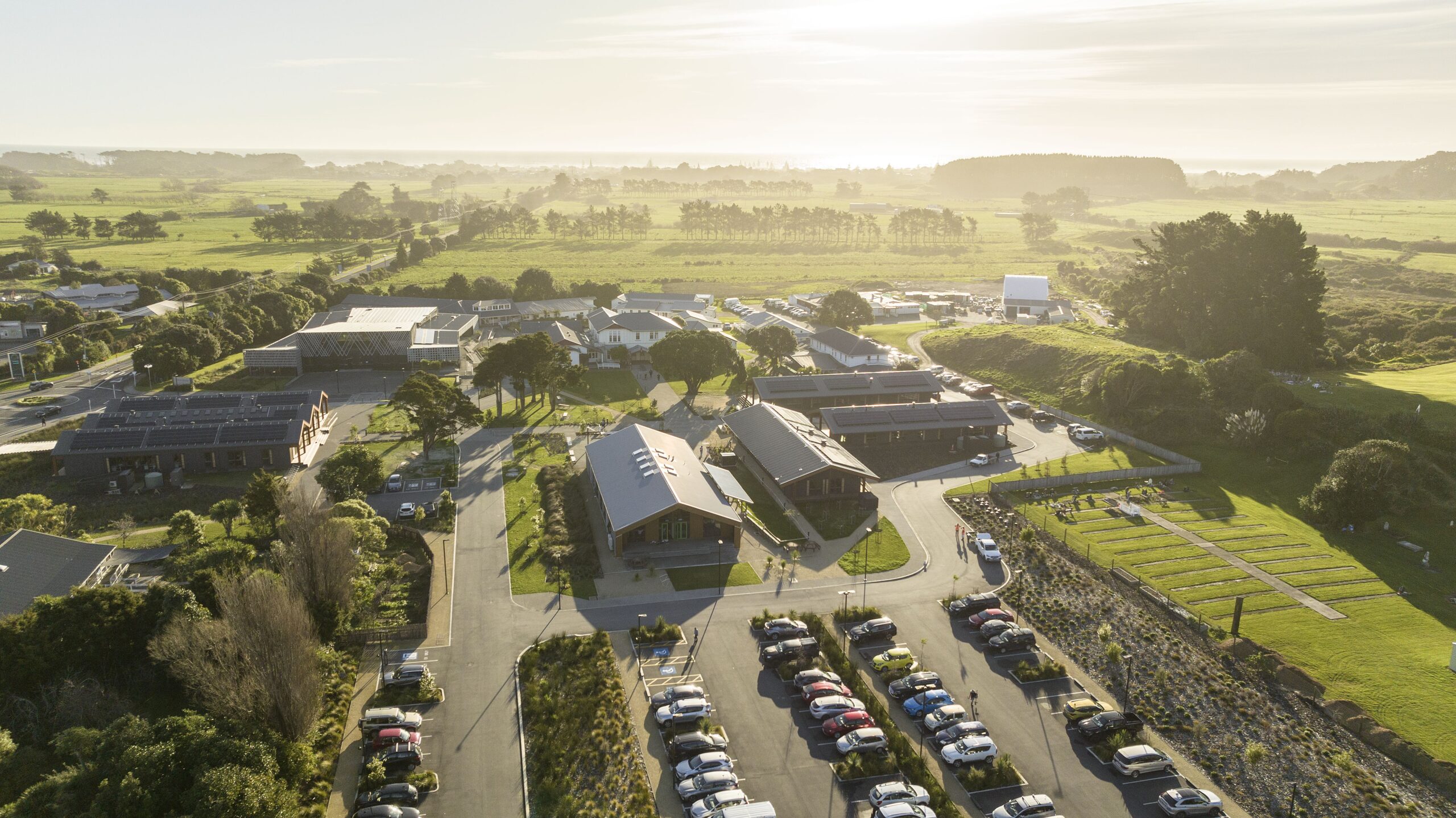 Aerial view of a college campus with modern buildings, arranged parking lots, and green lawns, set against open fields and distant coastline.