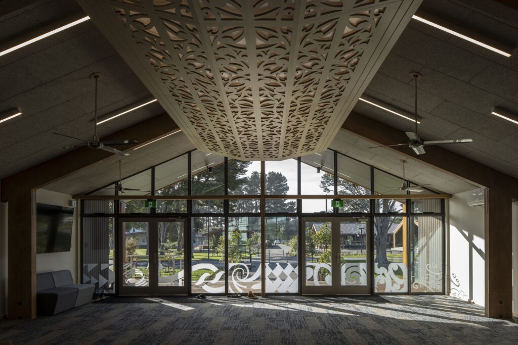 Interior view of a spacious lobby with a decorative perforated ceiling panel suspended overhead and large glass doors with frosted patterns outside.