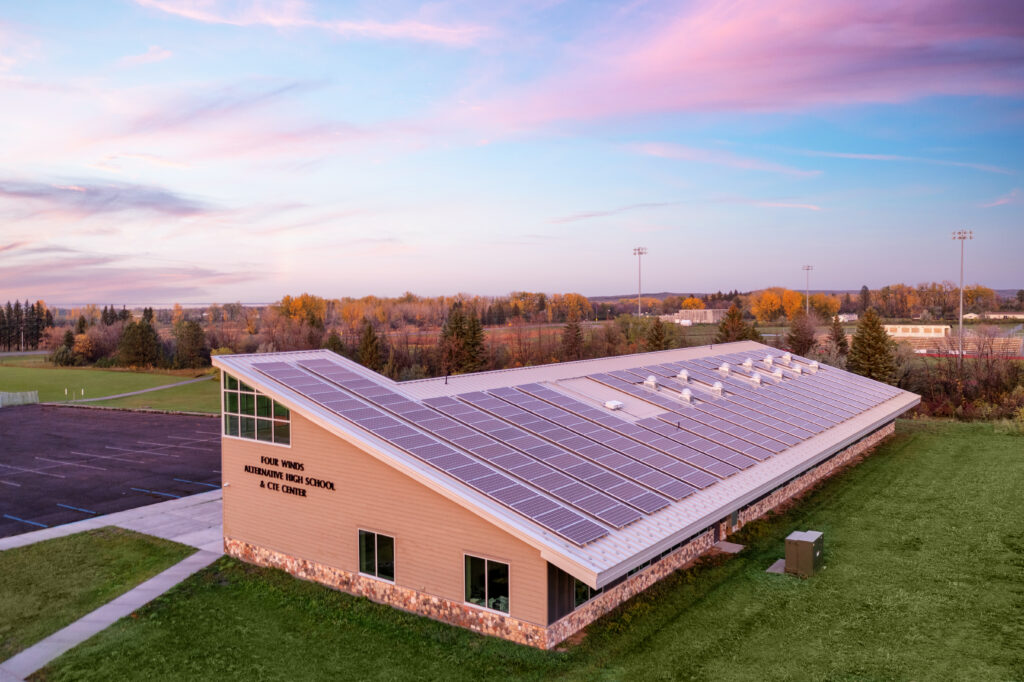 Aerial view of a sunlit school building with a solar panel roof and green grounds, with distant trees and stadium lights under a pink-blue sunset.