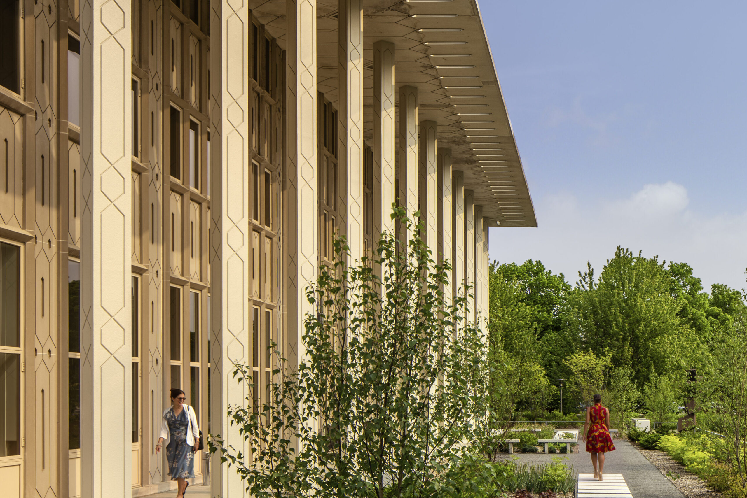 Modern beige campus building with tall decorative columns on the left, a pedestrian path, and greenery under a blue sky with trees in the background.