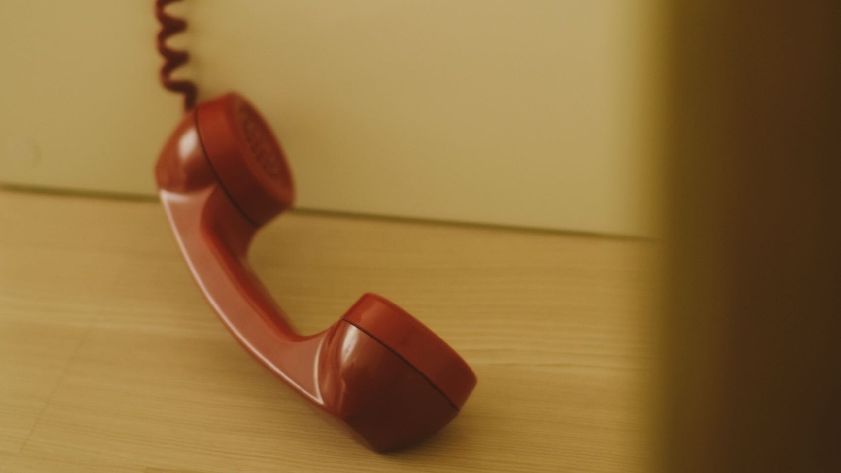 Red landline phone handset with a coiled cord hanging against a pale yellow wall surface.