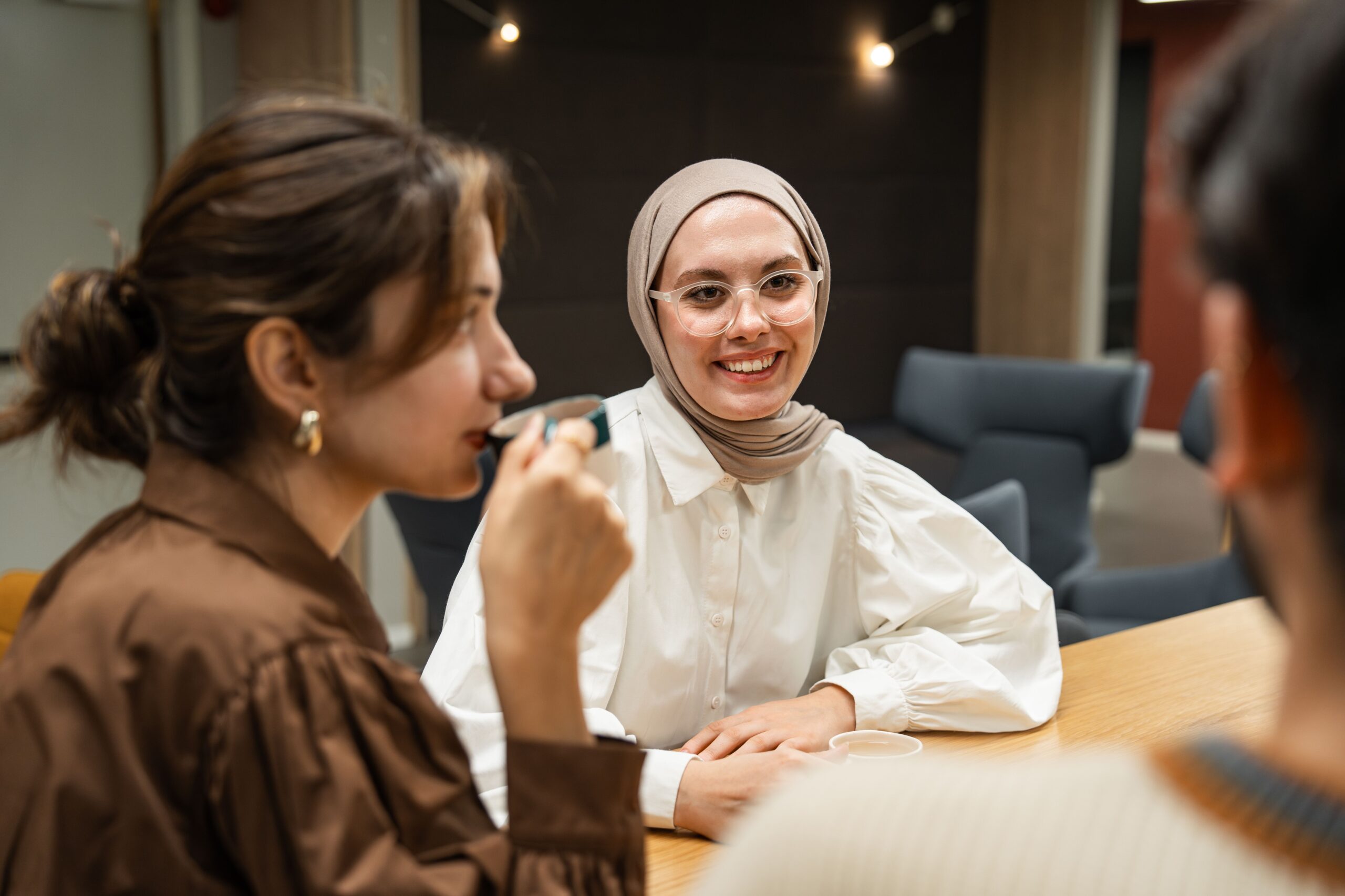Woman in a beige hijab and glasses smiles while chatting with colleagues at a wooden table in a modern meeting room.
