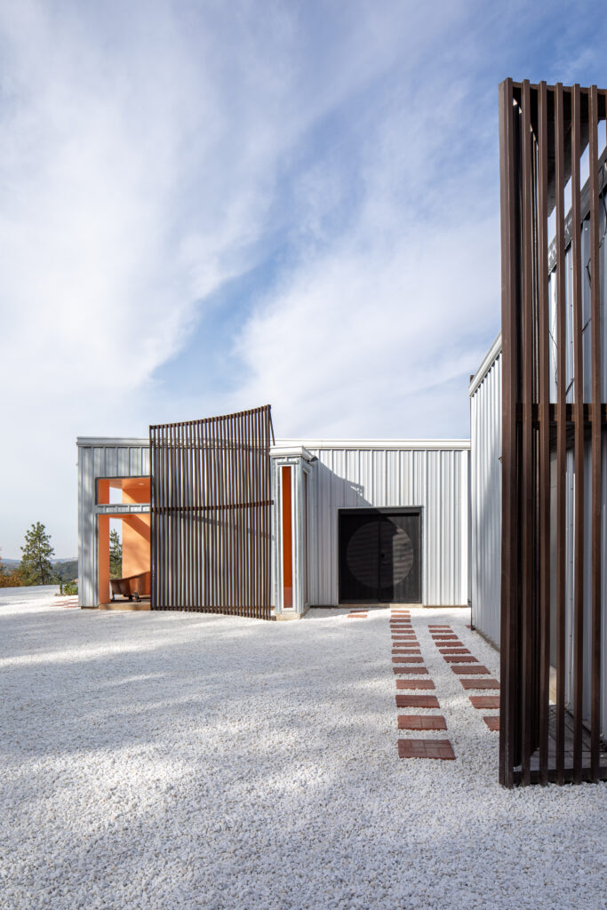 Modern building facade with vertical brown slats, orange-framed entryways, and a gravel yard under a blue sky.